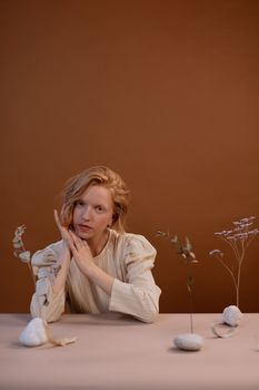 A serene portrait of a woman posing indoors with minimalist floral decoration against a brown background.