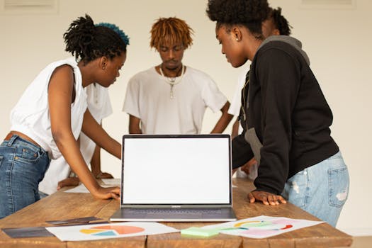 Group of young adults working together in an office, focused on a laptop with mockup screen.