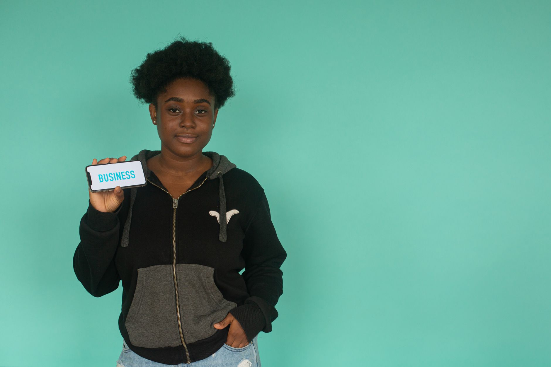 Confident woman in hoodie holding smartphone displaying 'business' against teal background.