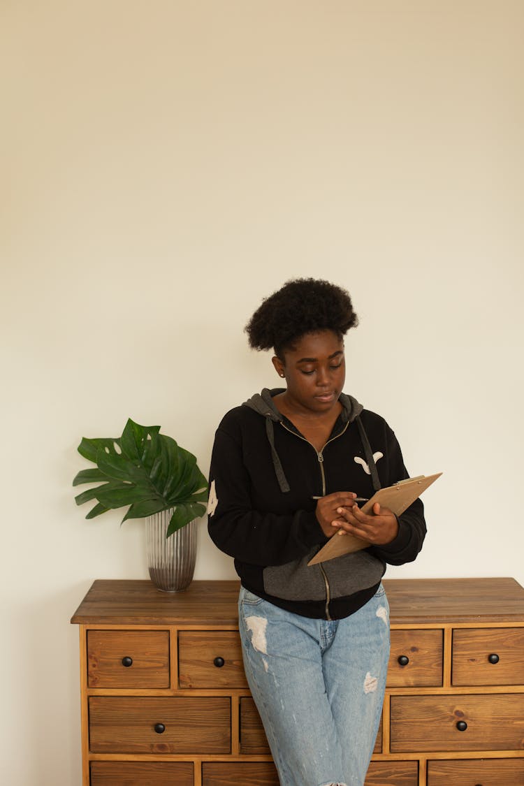 Woman With Afro Hairstyle Leaning Against Wooden Cupboard And Writing On Notepad