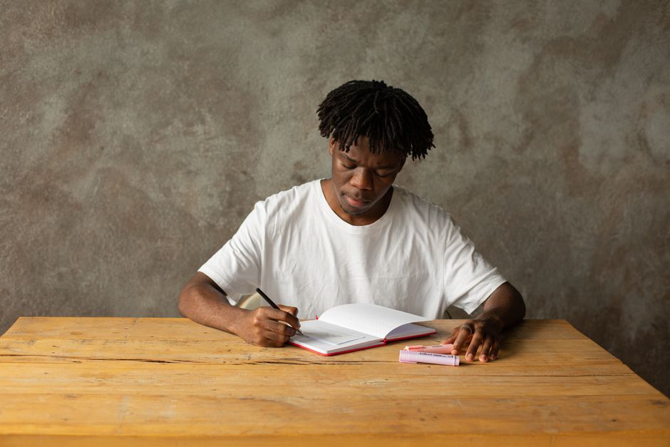 A young man in a white t-shirt focuses on studying at a wooden table indoors. A young man in a white t-shirt focuses on studying at a wooden table indoors.