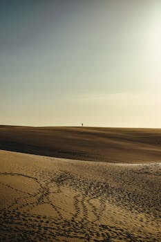 A lone figure stands on the vast sand dunes of Barreirinhas, Brazil, during sunset.