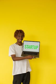 Portrait of young man holding a laptop displaying 'Startup' against a bright yellow background.
