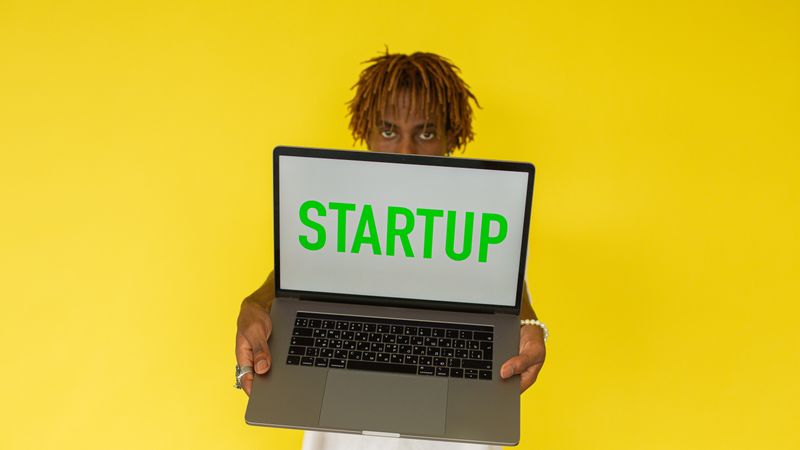 A young black man holds a laptop displaying 'Startup' against a vibrant yellow background.