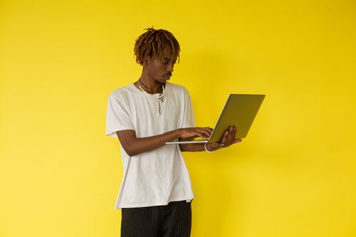 Free Young African man focused on his laptop against a bright yellow background, represents modern work culture. Stock Photo