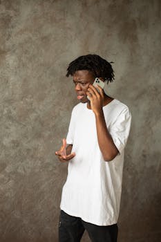 Young man in a white t-shirt engaged in a phone conversation indoors, showing an expressive gesture.