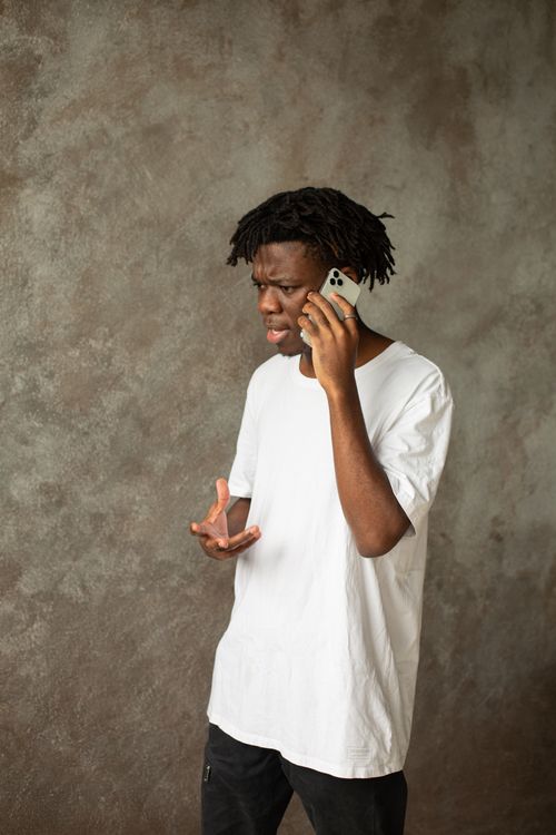 Free Young man in a white t-shirt engaged in a phone conversation indoors, showing an expressive gesture. Stock Photo