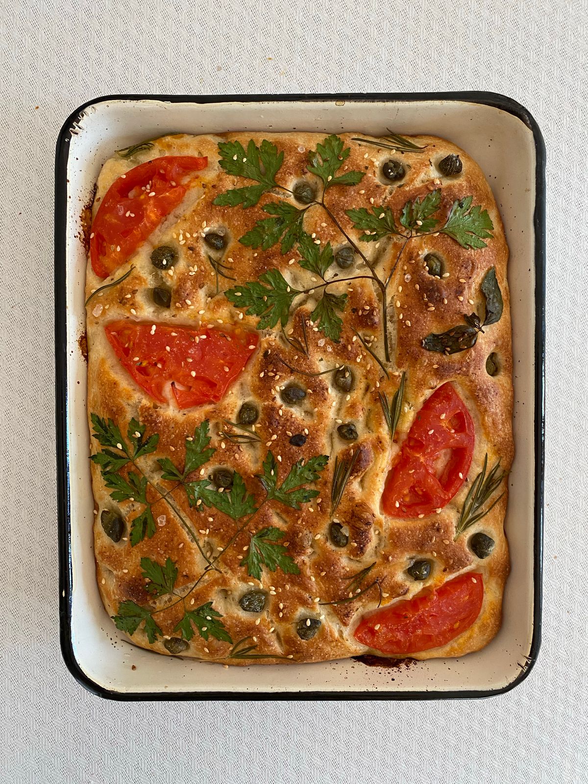 Homemade focaccia topped with fresh herbs and tomatoes in a baking tray.