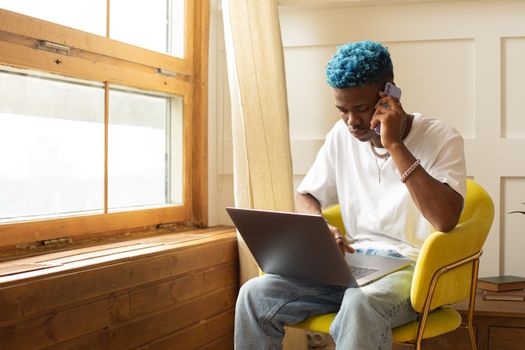 African American man working on laptop and phone call indoors with bright window lighting.