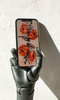 A black robotic hand holding a smartphone displaying a floral wallpaper, casting a shadow on a light wall.