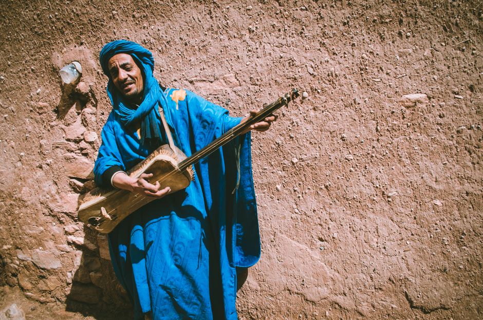 A Moroccan musician in vibrant blue plays a traditional instrument against a rustic wall.