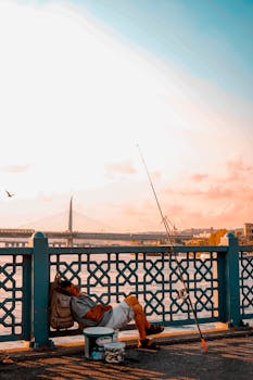 A man rests by a river, fishing rod in hand, in a serene sunset setting.