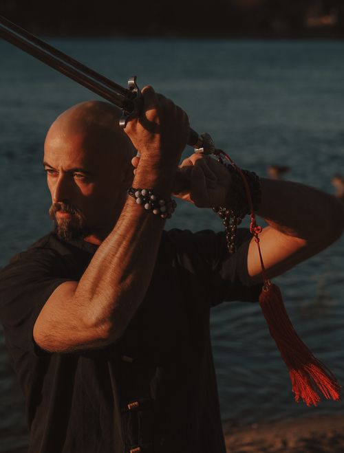 Free A bald martial artist poses dramatically by the water, holding a sword with a red tassel. Stock Photo