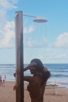 A woman enjoys a refreshing shower on a sunny beach day with ocean waves in the background.