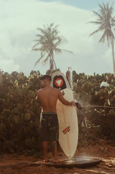 Shirtless man rinses surfboard in sunny tropical setting with palm trees.