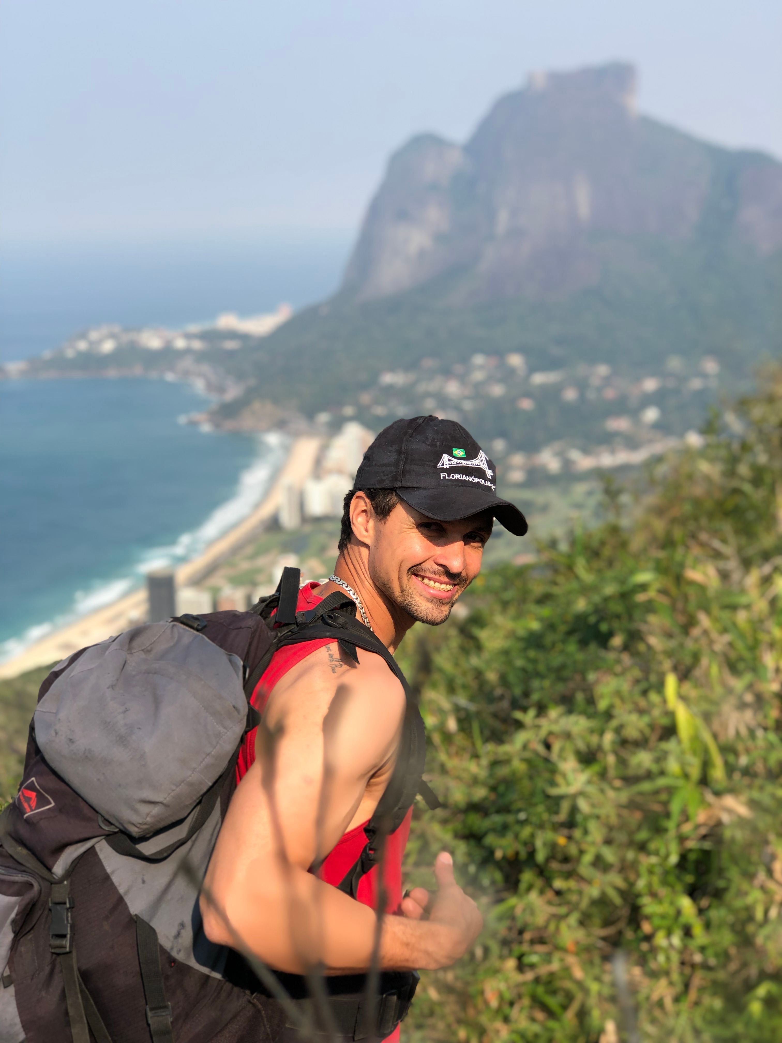 Smiling man hiking with a backpack overlooking Rio de Janeiro's picturesque mountains and coastline.