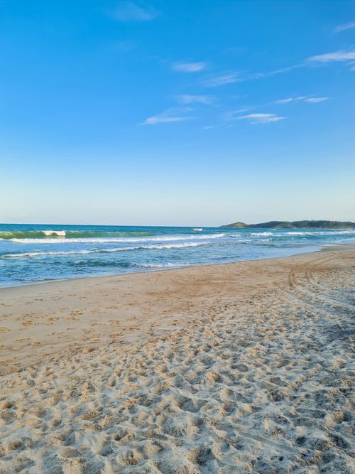 Free Serene sandy beach with gentle waves and a bright blue sky, perfect for relaxation and coastal exploration. Stock Photo