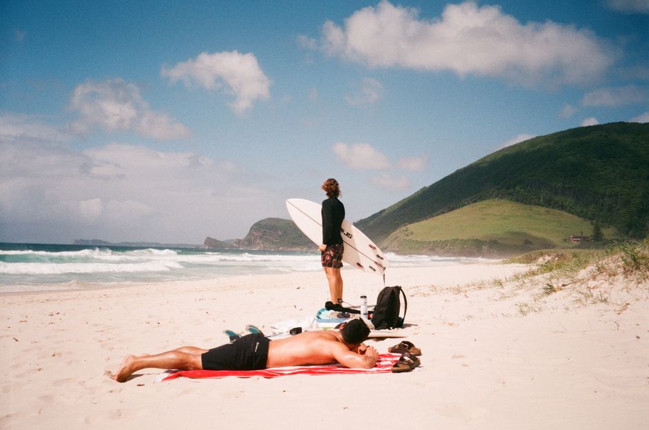 A surfer standing by the ocean with a surfboard while a man sunbathes on the sand.