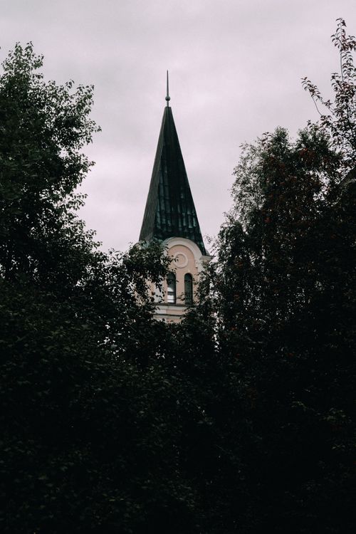 Free A gothic church spire peeks through dense foliage under a cloudy sky, capturing a moody atmosphere. Stock Photo