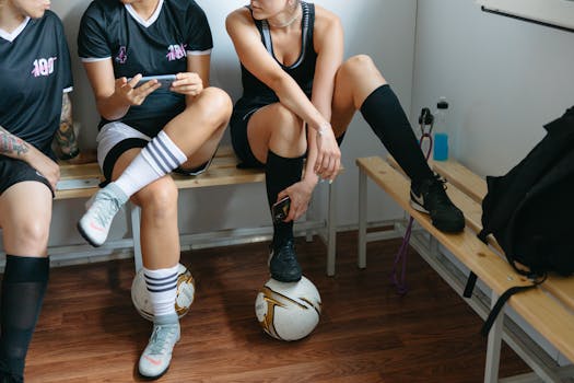 Three female soccer players sitting in a locker room, wearing sportswear and holding a phone