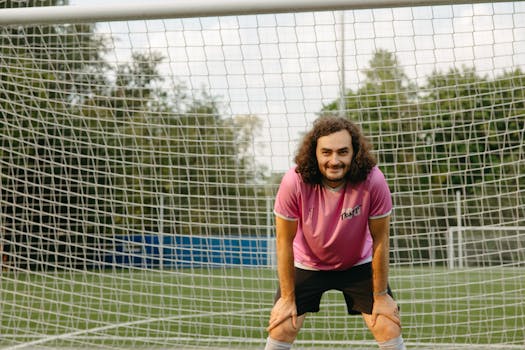 Smiling bearded athlete in pink soccer jersey posing in front of a goal on an outdoor field.