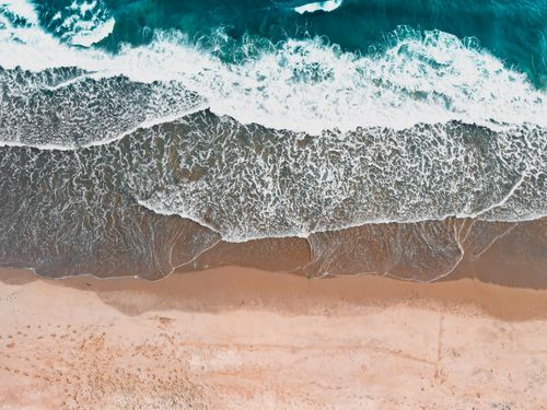 Free Drone view of ocean waves crashing onto a pristine sandy beach, creating stunning patterns. Stock Photo