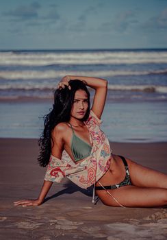 Young woman in a bikini sitting on a sandy beach, exuding relaxation and glamour.