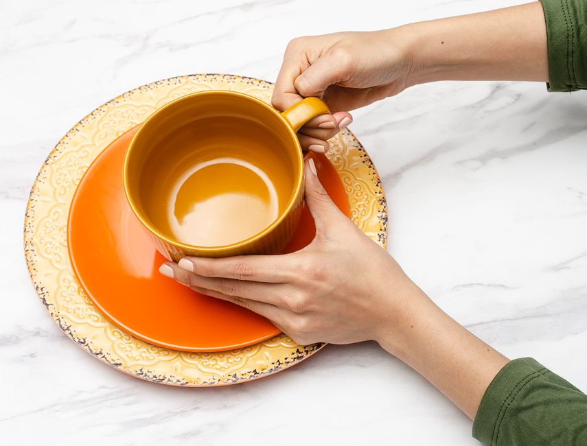 Hands holding a vibrant orange mug on a decorative saucer, placed on a white marble surface.
