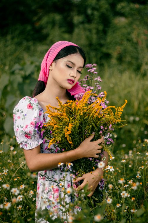 Free A young woman in a floral dress holding a bouquet, eyes closed in a serene field setting. Stock Photo