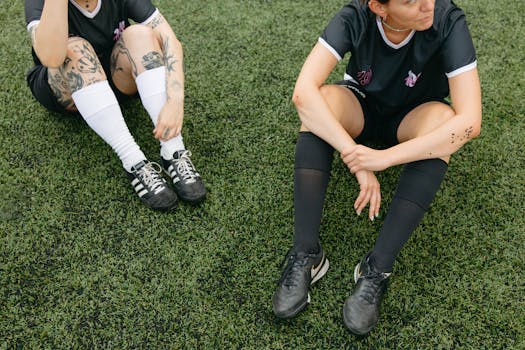 Two female soccer players sitting on a grass field in uniforms, taking a break.