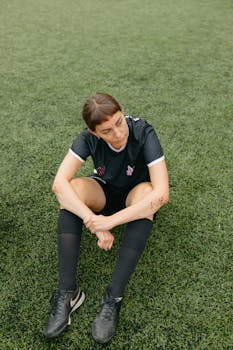 A woman in a football uniform takes a break sitting on the grass field.