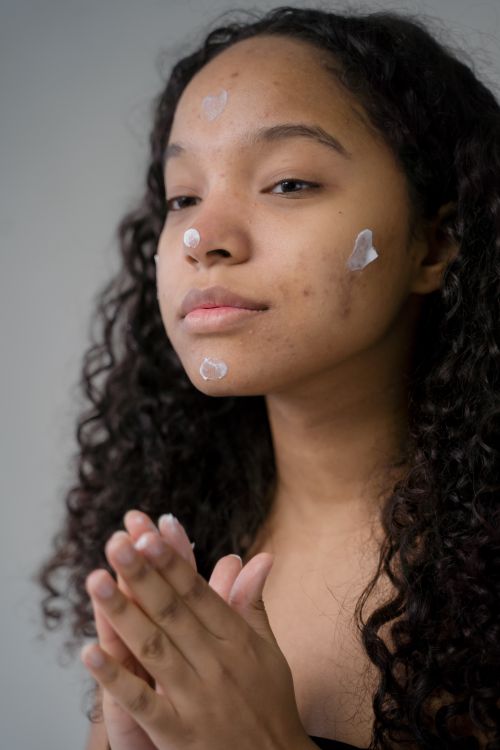 Free Young woman with curly hair applying skincare cream in a studio setting, focusing on beauty and hydration. Stock Photo
