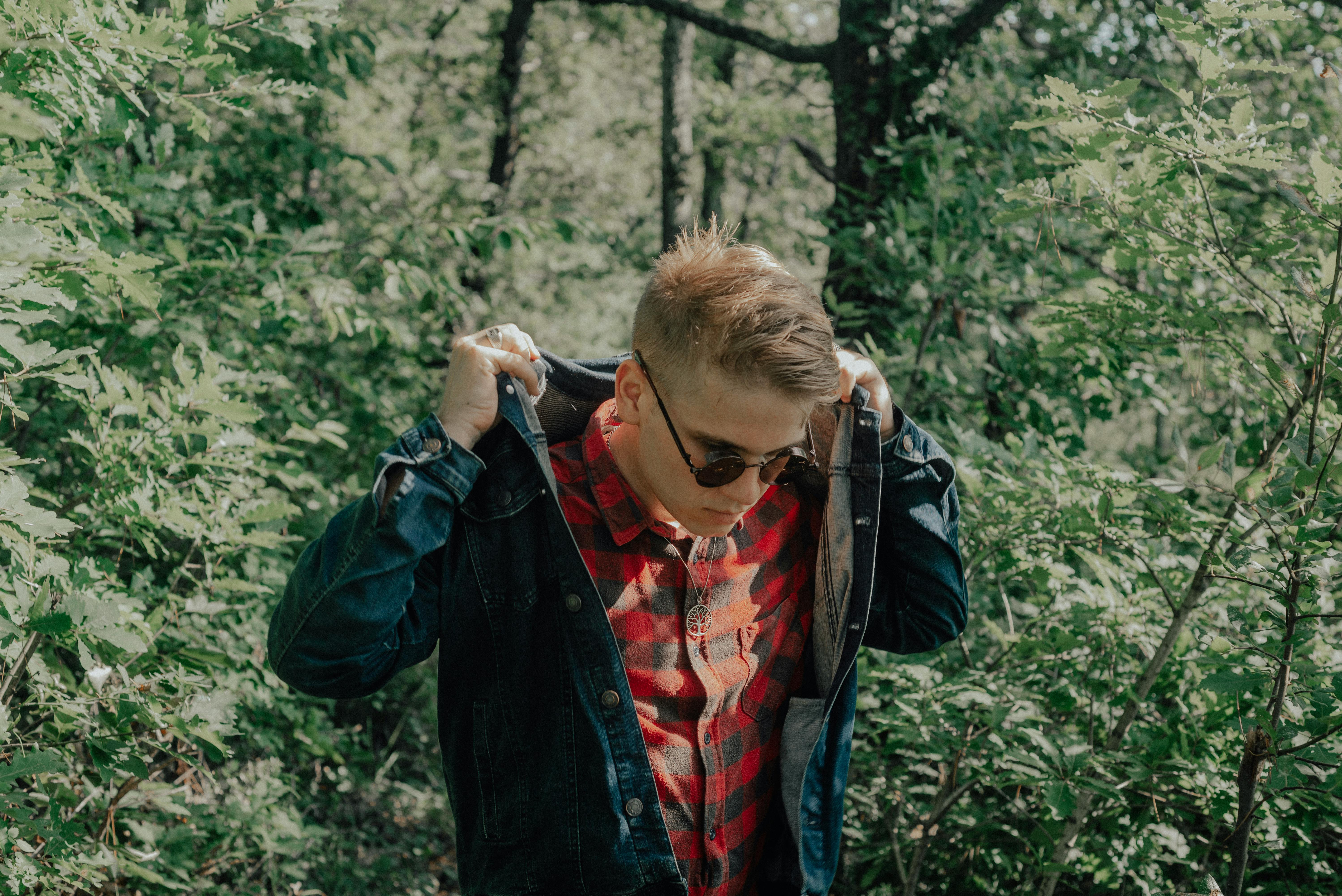 A young man adjusts his denim jacket while exploring a sunny forest.