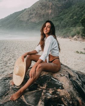 A woman enjoying a sunny day on a scenic Rio de Janeiro beach, sitting on a rock with a hat.