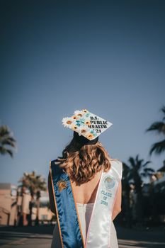 A young woman celebrates her graduation outdoors under a clear sky with palm trees.