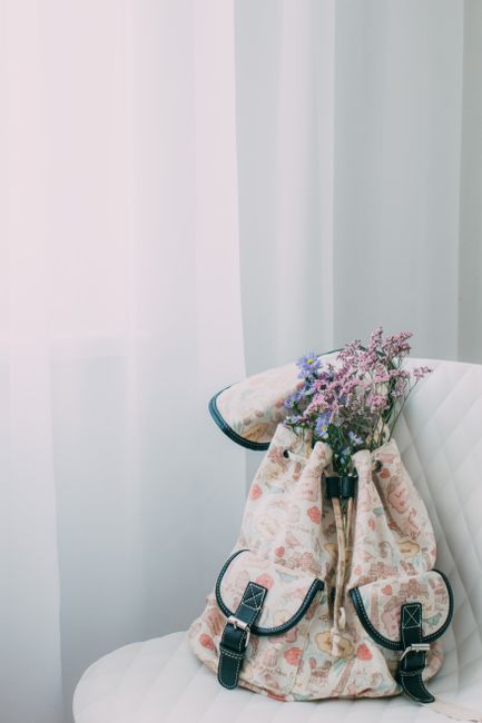 Patterned backpack filled with wildflowers on a white chair indoors, soft lighting.
