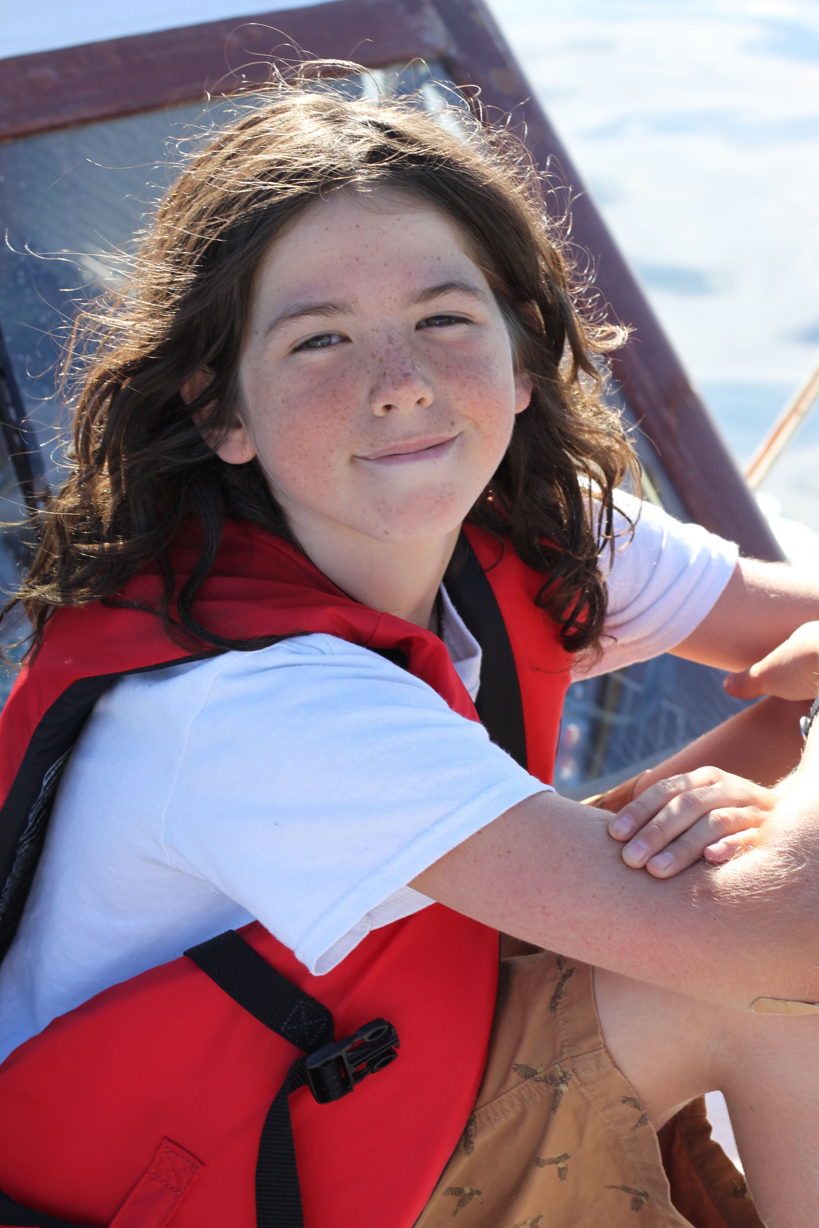 A smiling girl in a red life vest on a boat, enjoying a sunny day outdoors.