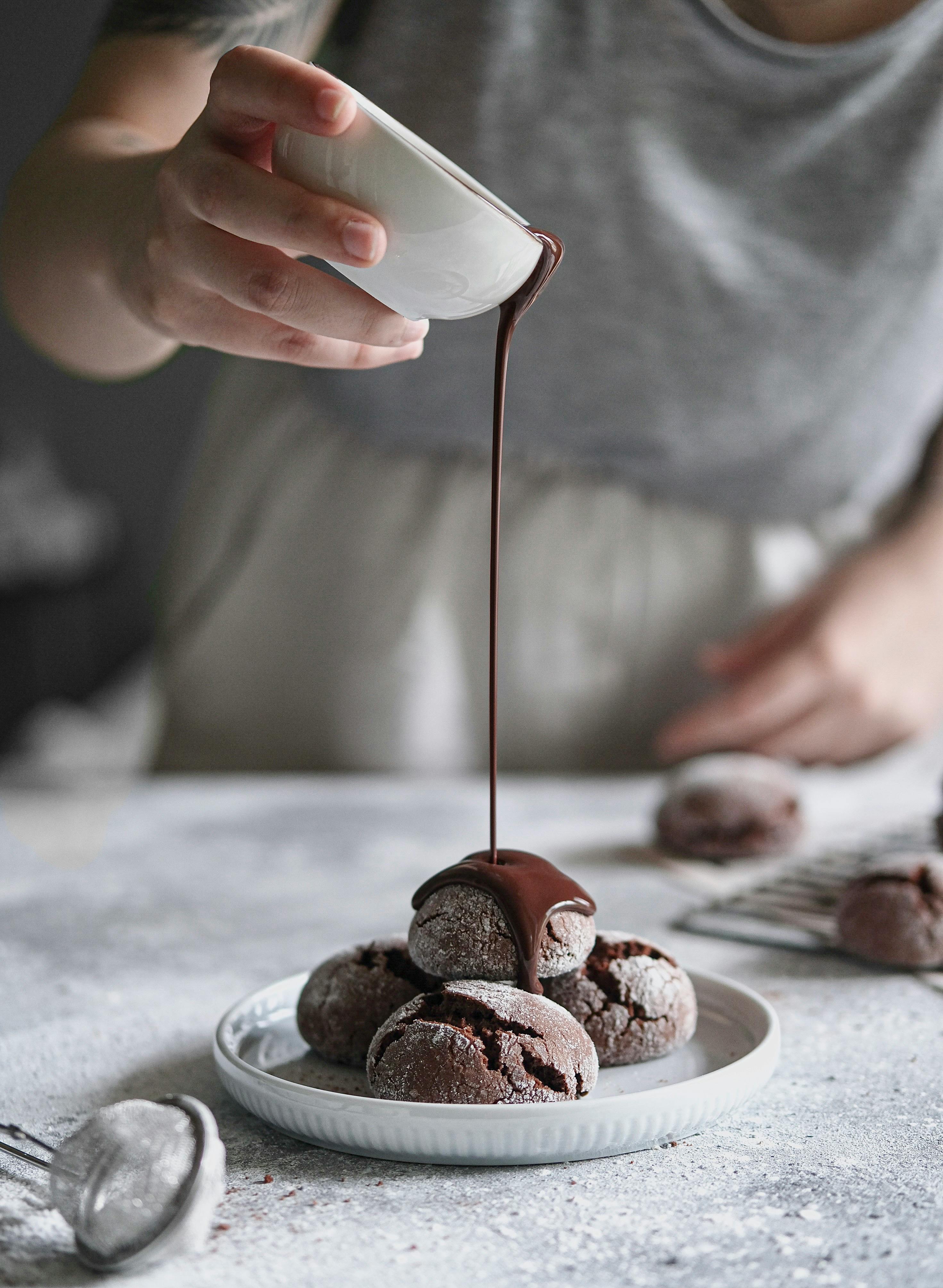 A person drizzles rich chocolate sauce over homemade chocolate crinkle cookies on a plate.