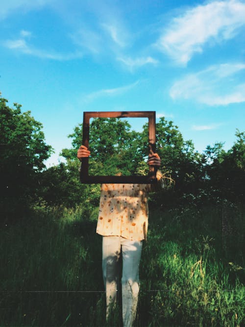 A Person in Yellow Button Up Shirt Standing on Green Grass Holding Brown Framed Mirror