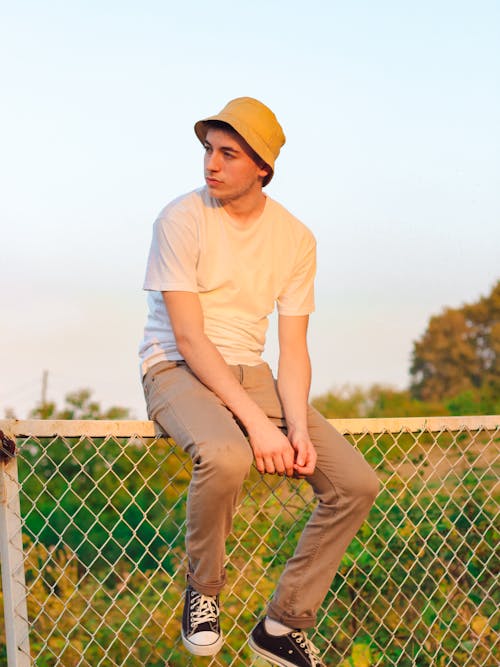 Photo of a Man in a Bucket Hat Sitting on Top of a Chain Fence