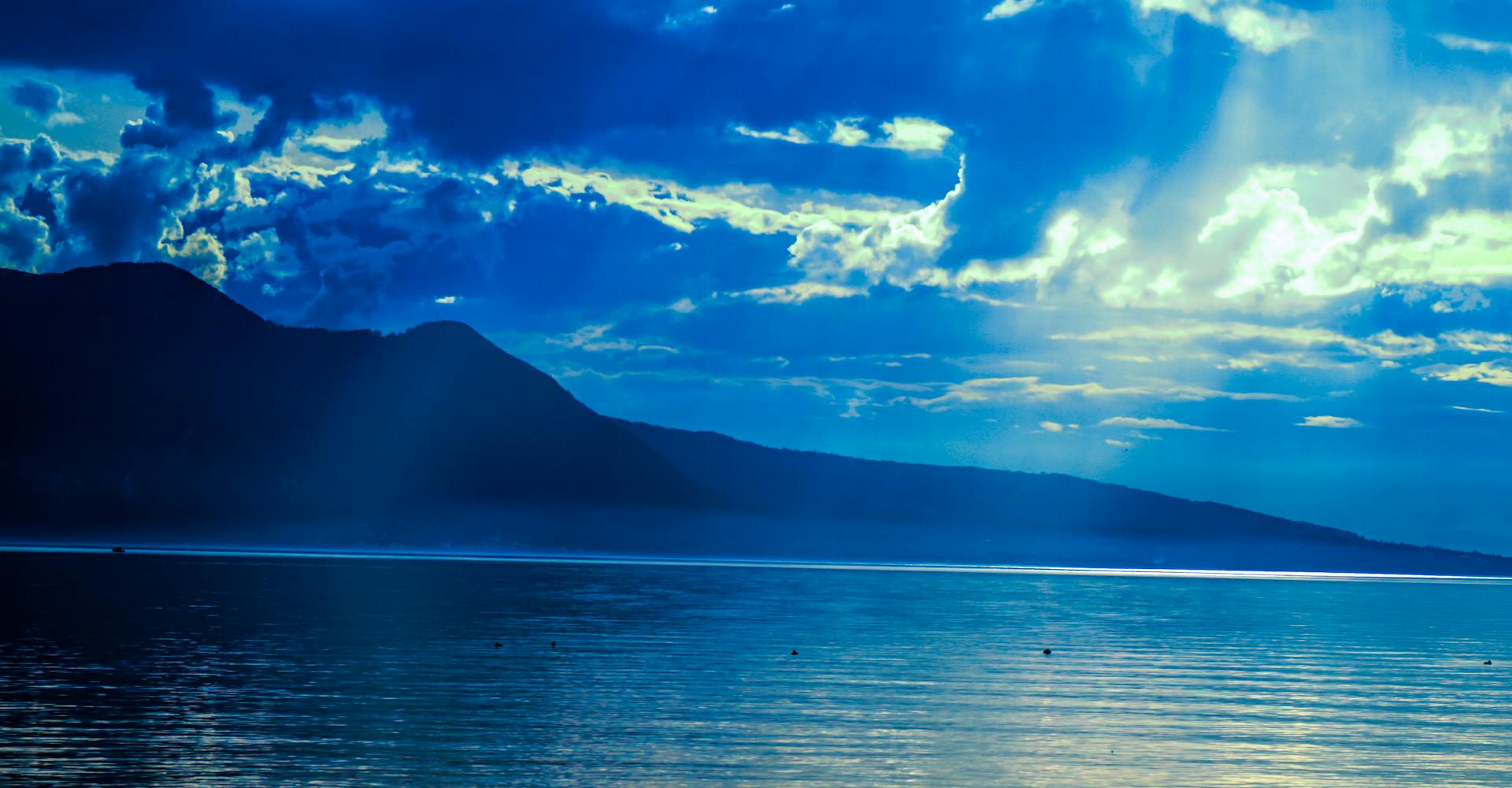 Captivating view of a serene sea with mountains and dramatic cloud formations under a vibrant blue sky.