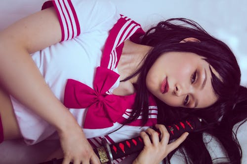 Woman in White and Red Crop Top With Bow Accent Holding Black and Red Handled Sword While Lying