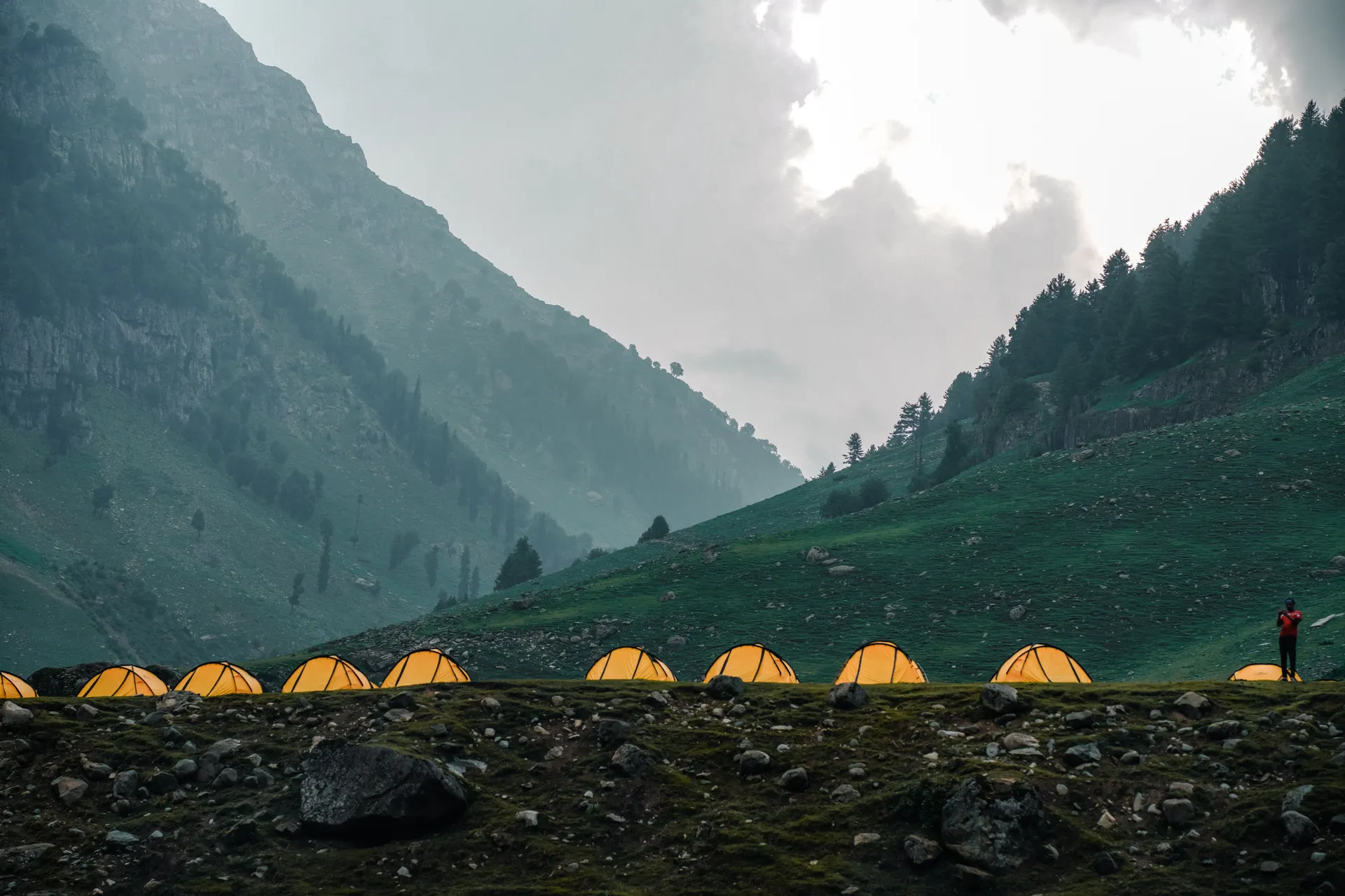 Scenic mountain campsite with illuminated tents and lone hiker under cloudy sky. Scenic mountain campsite with illuminated tents and lone hiker under cloudy sky.