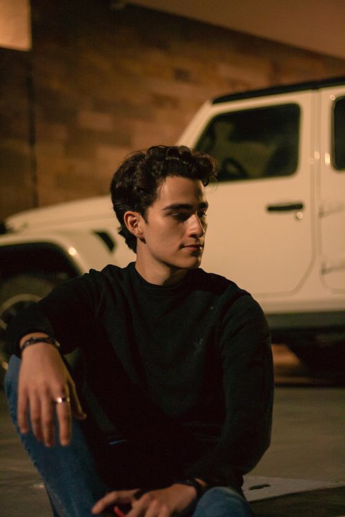 Free Man in black sweater sits by a white car in a dimly lit urban environment. Stock Photo