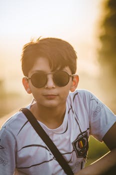 A fashionable young boy with sunglasses poses outdoors in warm lighting.