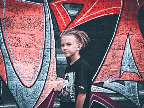 Photo of a Boy in a Black Crew Neck Shirt Standing Near a Wall with Graffiti