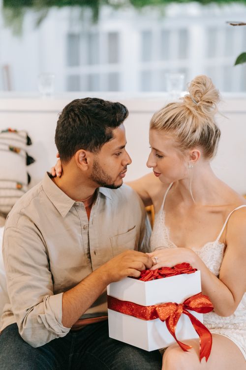 Free A young couple sharing a tender moment while exchanging a gift in a cozy indoor setting. Stock Photo