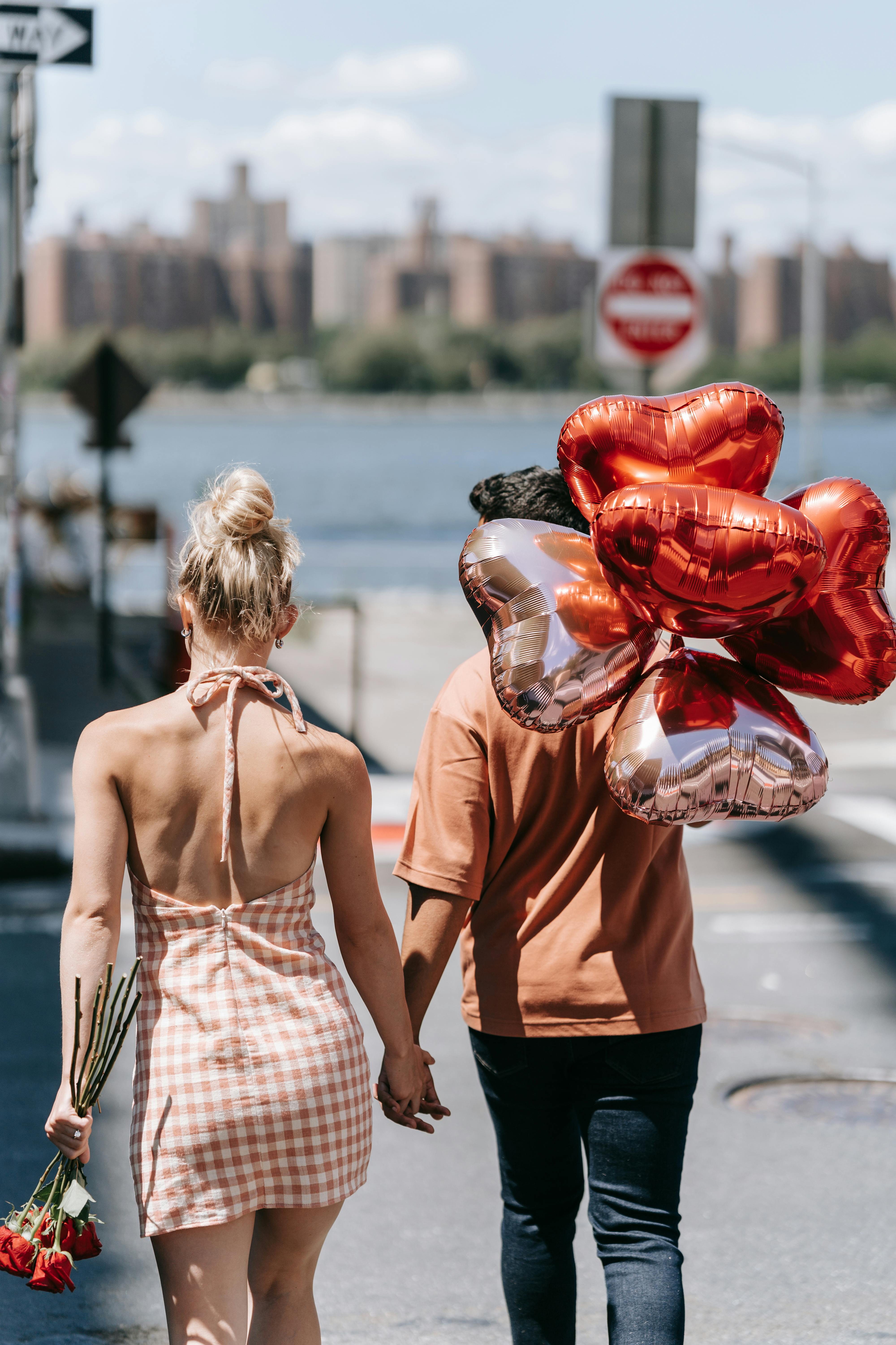 A couple holding hands with heart-shaped balloons and roses, walking in an urban setting.