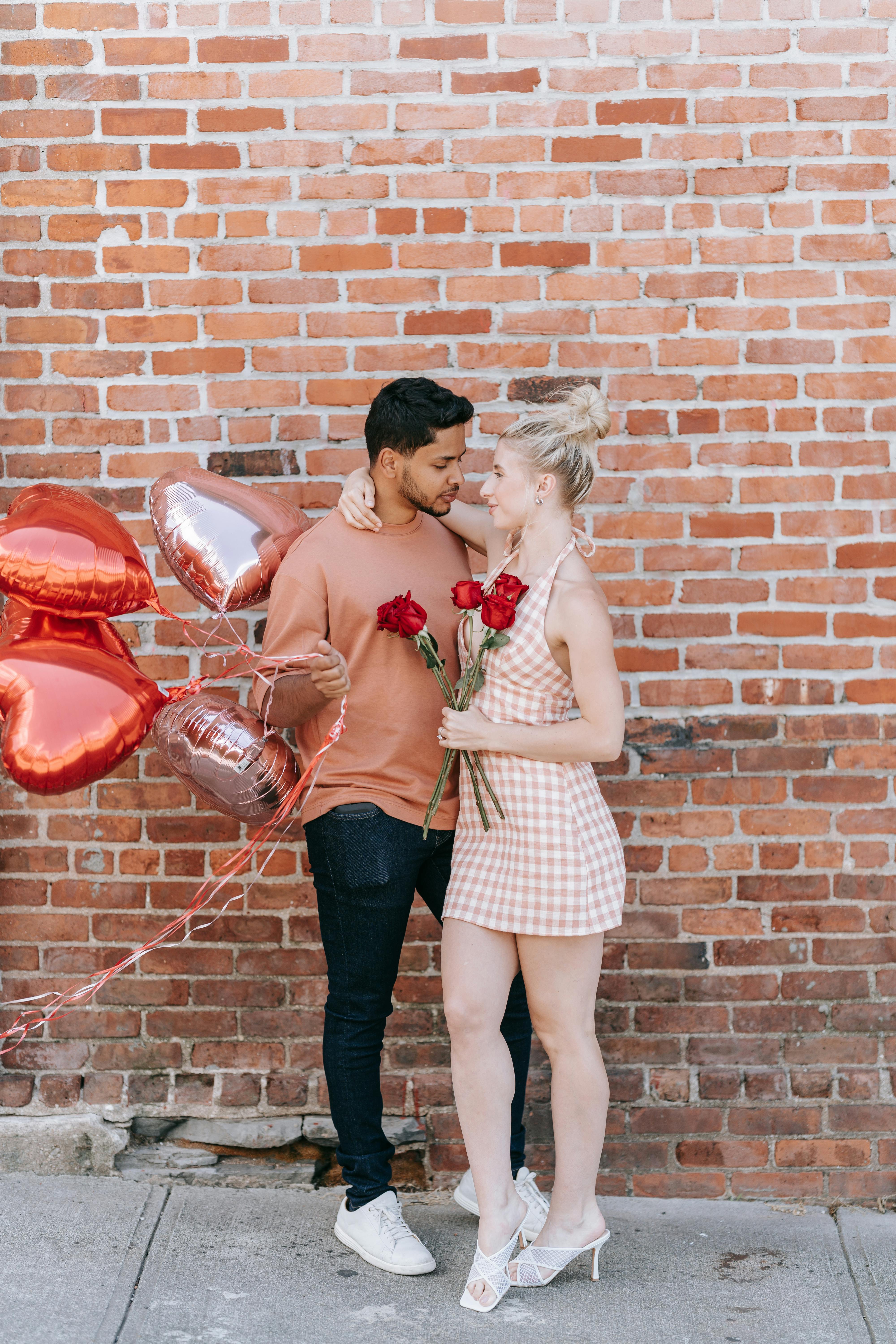 A couple sharing a romantic moment with roses and heart-shaped balloons against a brick wall.
