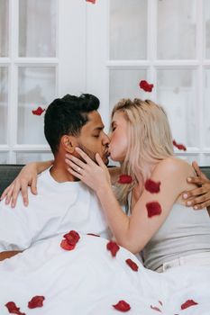 A loving interracial couple kissing amidst rose petals on a cozy bed, capturing an intimate and romantic atmosphere.